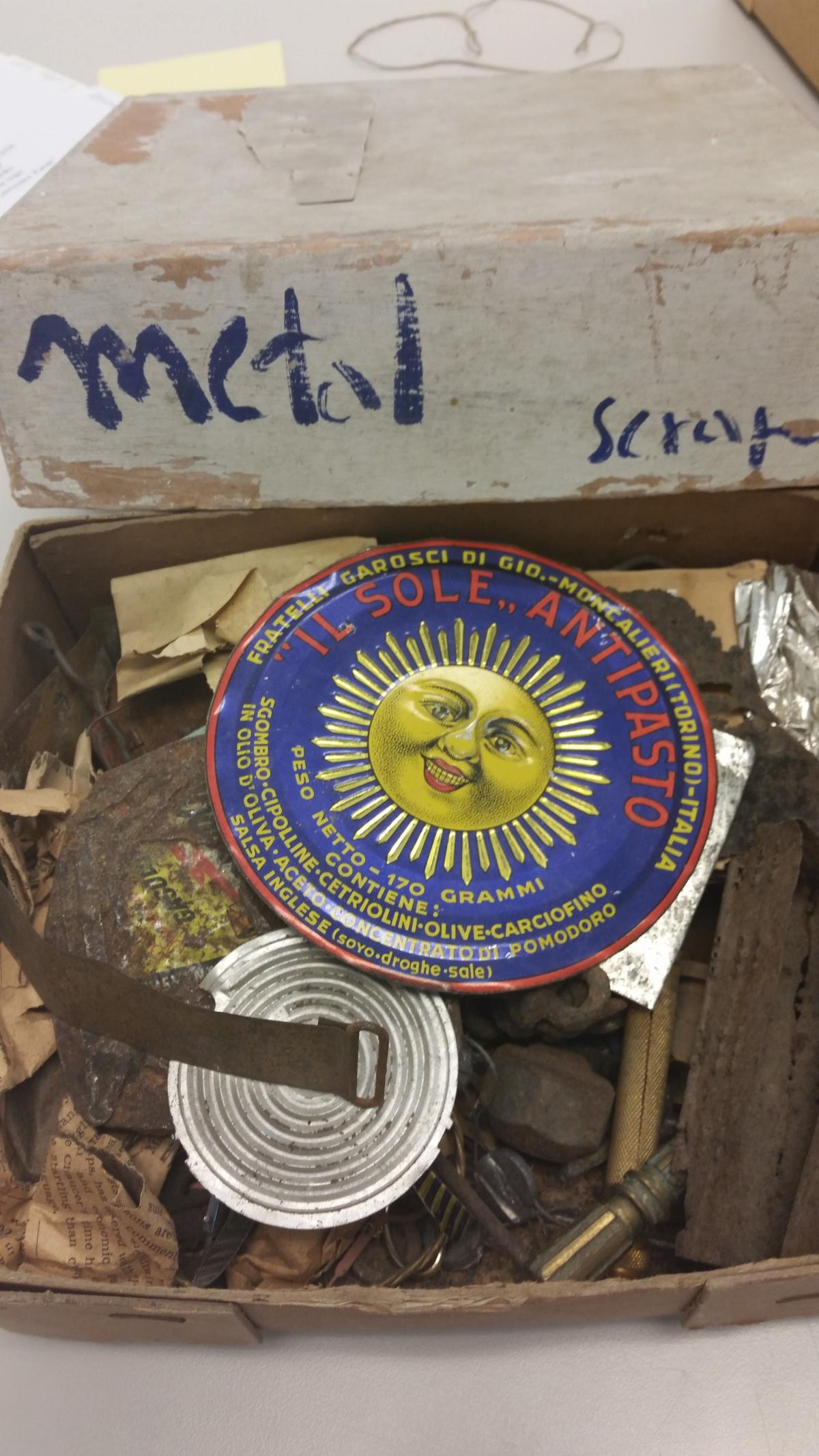 A box marked metal scapes in blue marker opened to an image from a jar of olives featuring a yellow sun; below scraps of metal