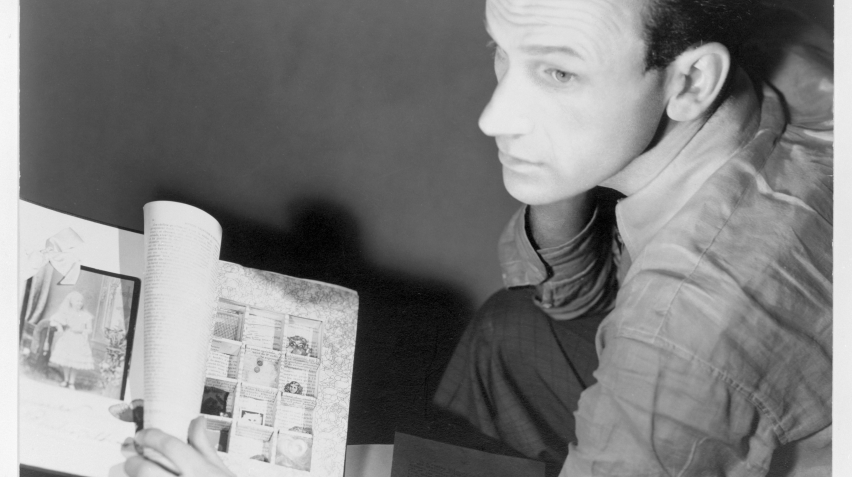 A black and white photo of a man sitting looking away from the camera, his artwork in front of him