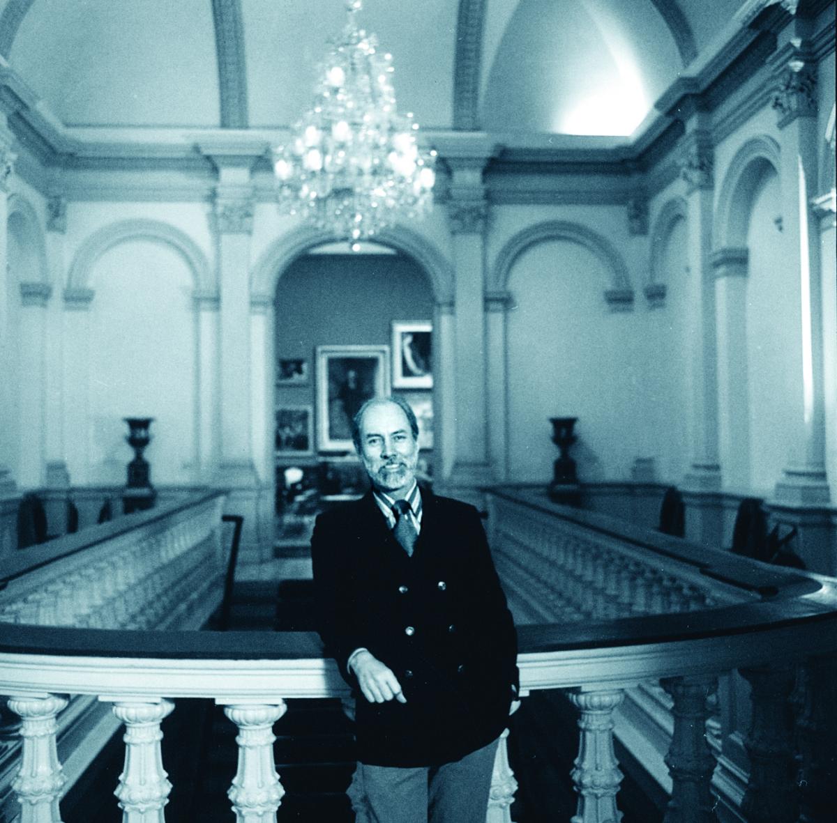 A white man wearing a dark blazer stands leaning on a balustrade. He has a slight smile.