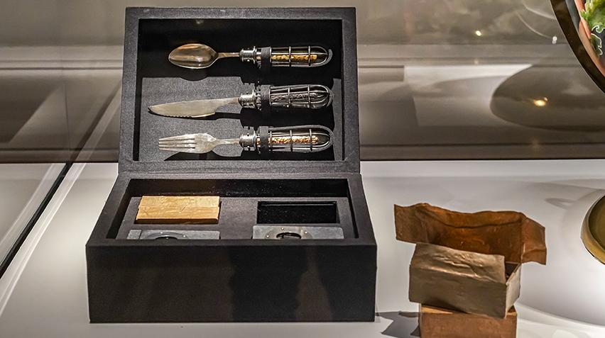 A black case that is open to reveal smaller compartments and utensils mounted in the lid. Boxes wrapped in brown paper are stacked on the side.