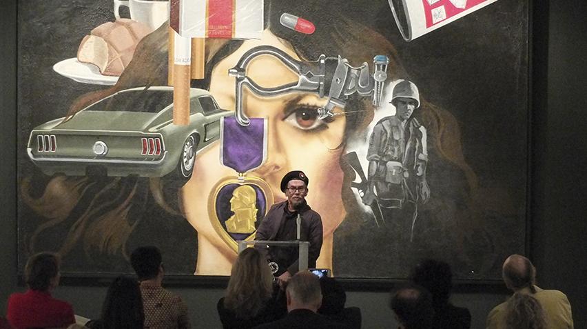 A man wearing a beret stands in front of an artwork. He is speaking to an audience.