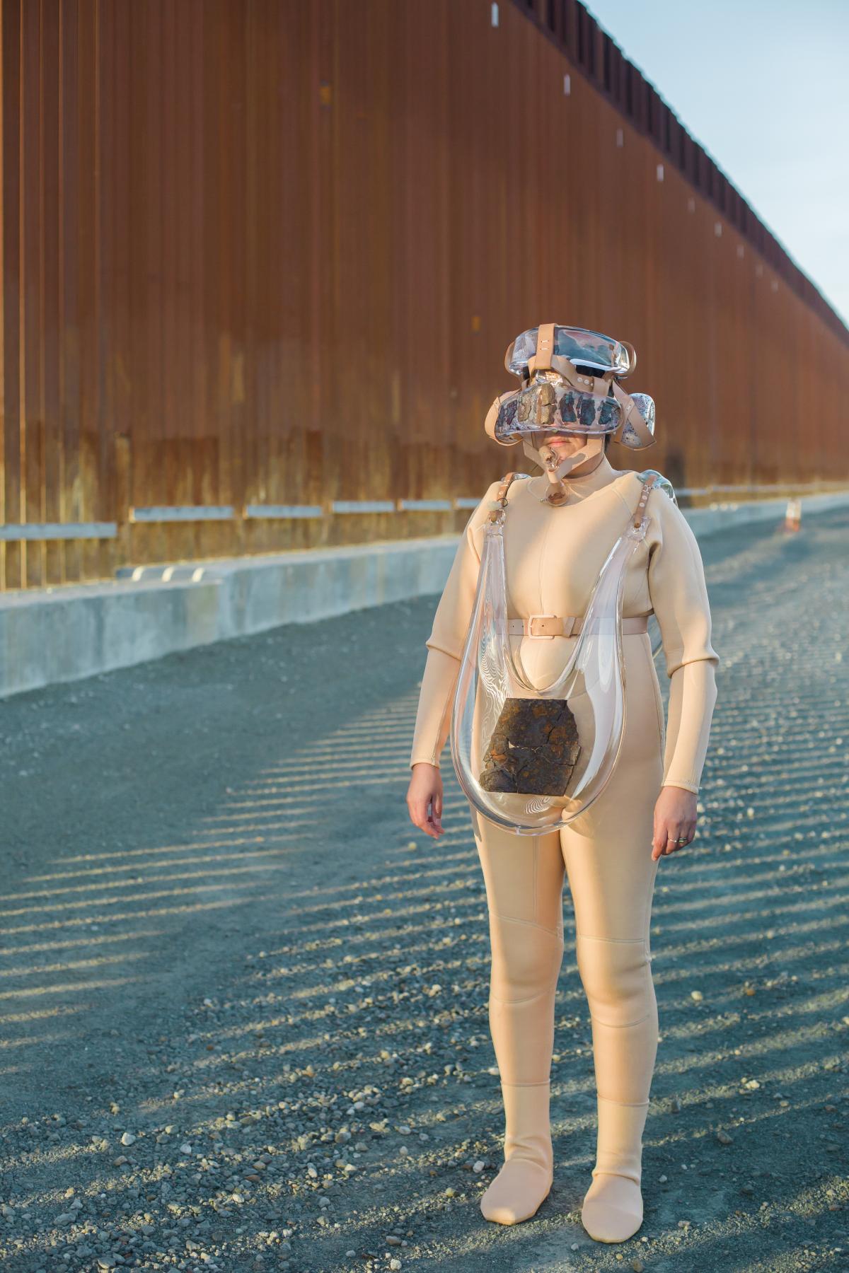 Artist Tanya Aguiñiga wearing a suit made of glass elements while standing next to the U.S./Mexico border wall