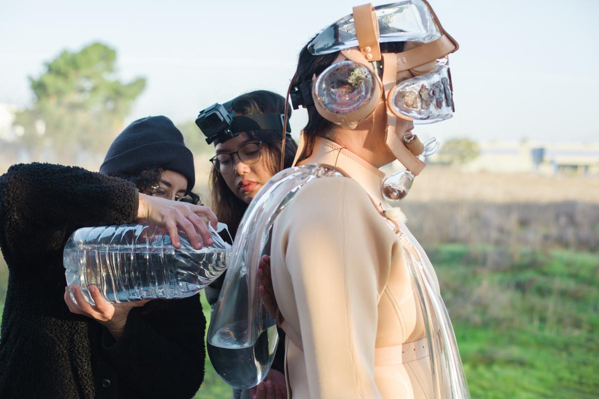 Two Latina women pour water into the glass elements of artist Tanya Aguiñiga's suit.