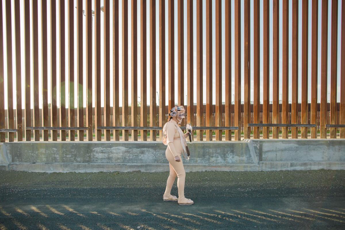 Artist Tanya Aguiñiga wearing a suit made of glass elements while walking next to the U.S./Mexico border wall