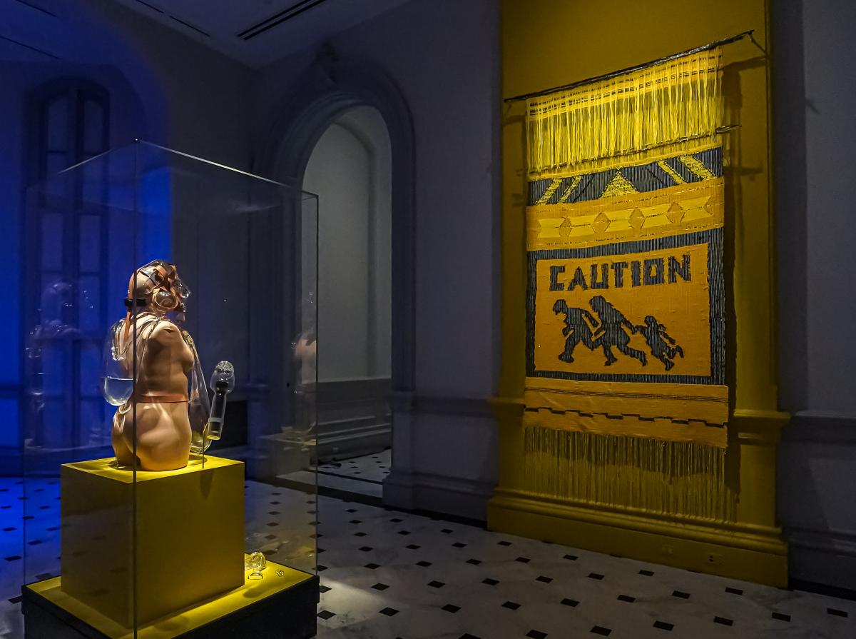 Two artworks installed in a dark gallery. A sculpture faces a bright yellow wall hanging with the the word "Caution" woven into it.