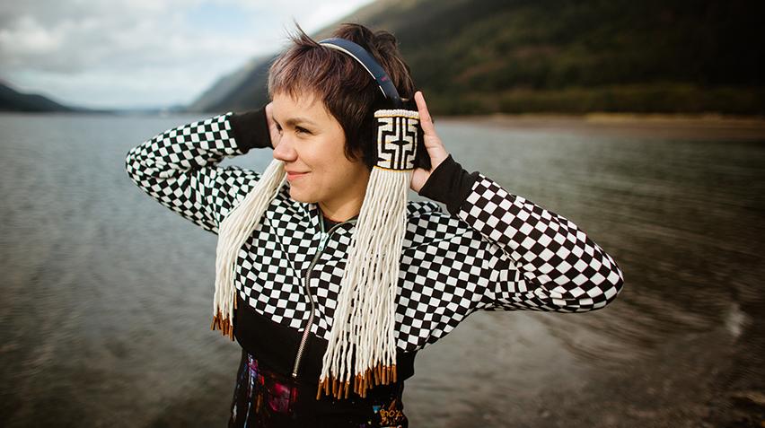 Blog - RI 23 Intro post, Memorial Beats, home page crop A person in a black and white checkered sweater stands holding headphones. They are also black and white, woven with an Indigenous motif and have long white fringe.