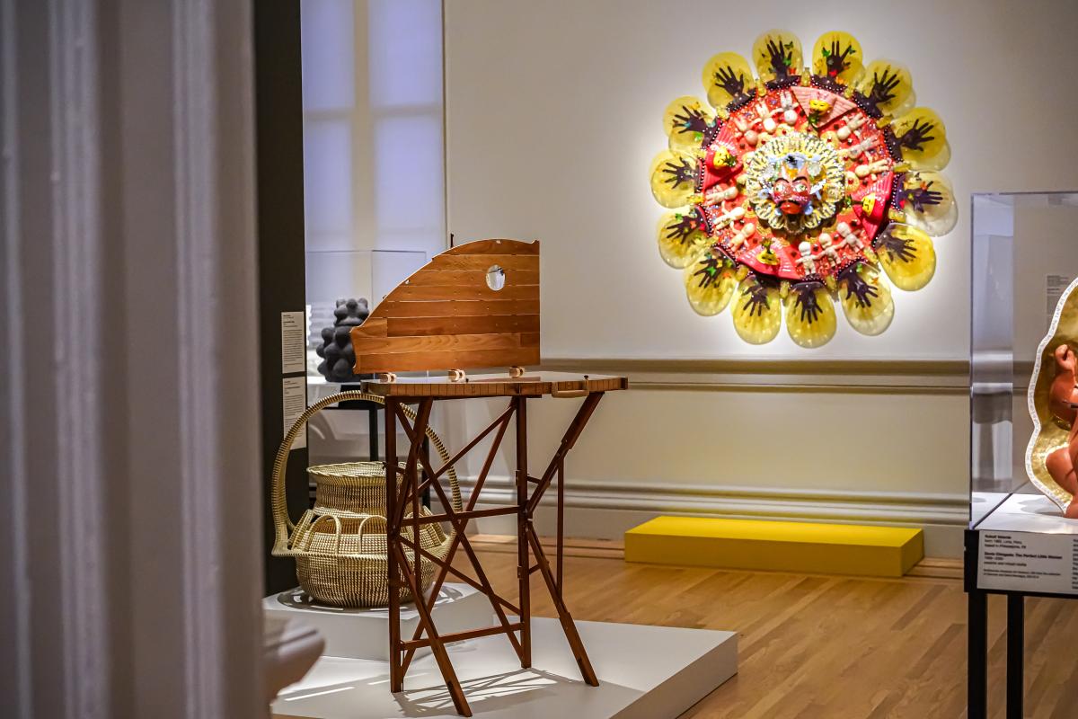 Installation view of a circular blown glass sculpture hanging on a gallery wall. There is an open wooden case on a stand in front of it.