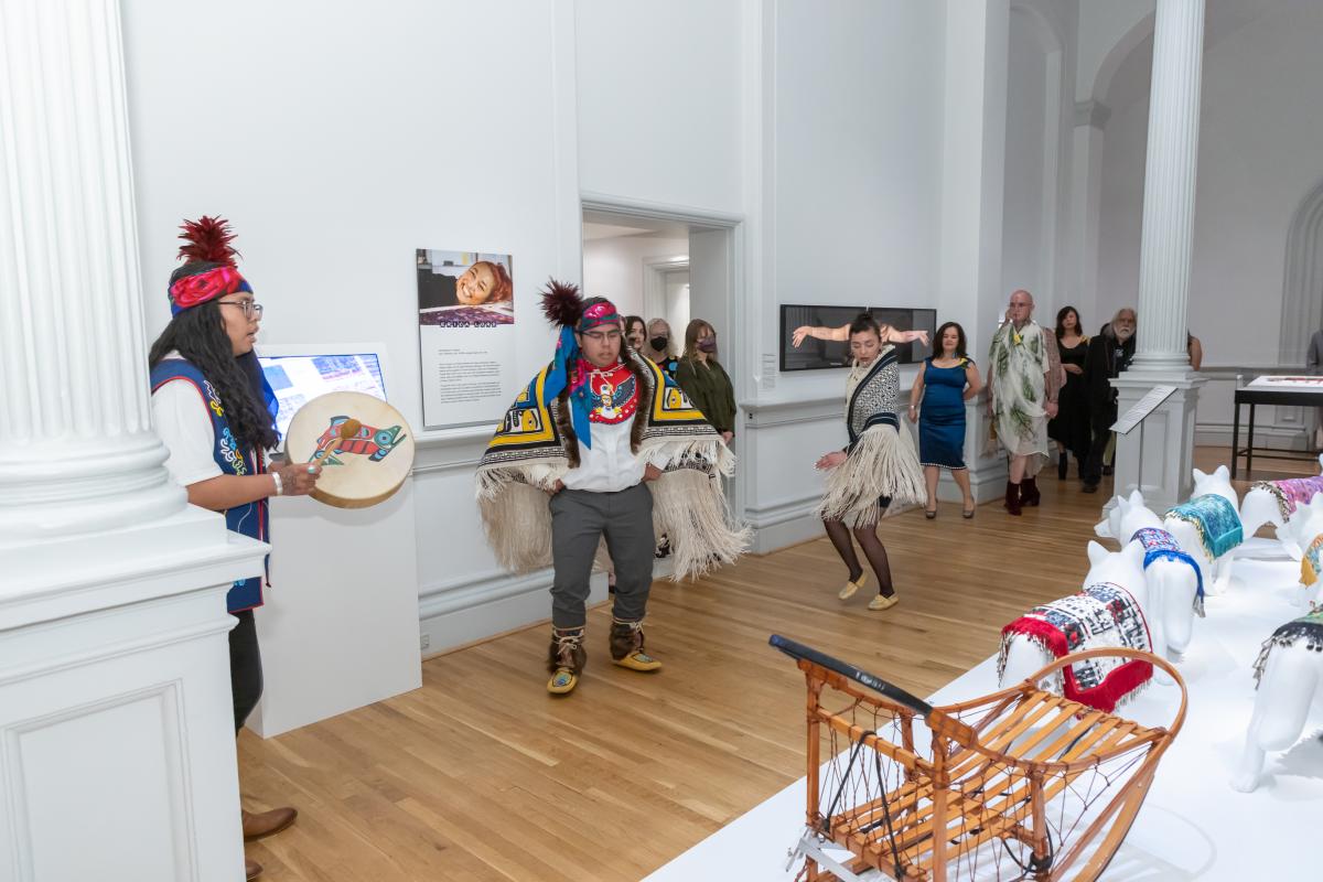 A performer wears a blanket with traditional Alaskan Native motifs. She dances in the museum gallery while Native drummers play beside her.