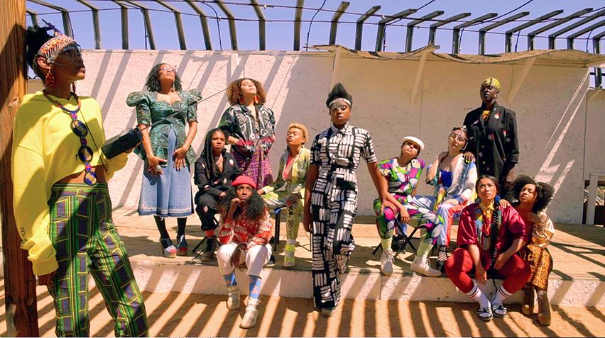 Blog - Afrofuturism, Cauleen Smith, Sojourner 2, home page crop A group of women standing outside under a slatted-canopy. The shadows of the slats cross the image.