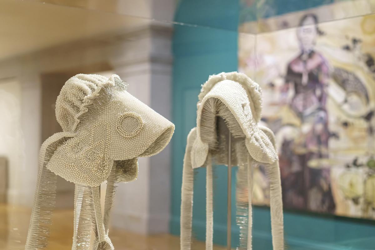 Two off-white, beaded bonnets on display in a glass case. In the background is a large painting.