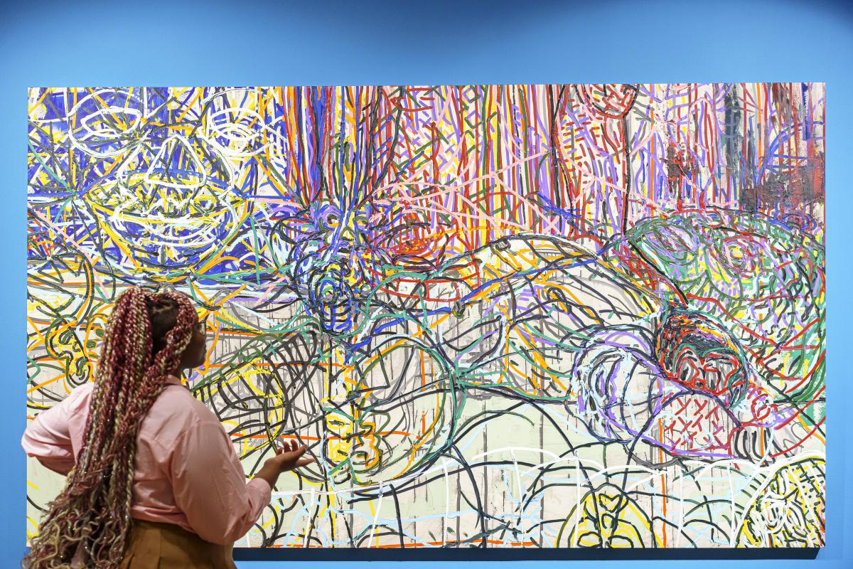 A woman with long braids is looking at a vibrant abstract painting, which hangs on a bright blue wall.