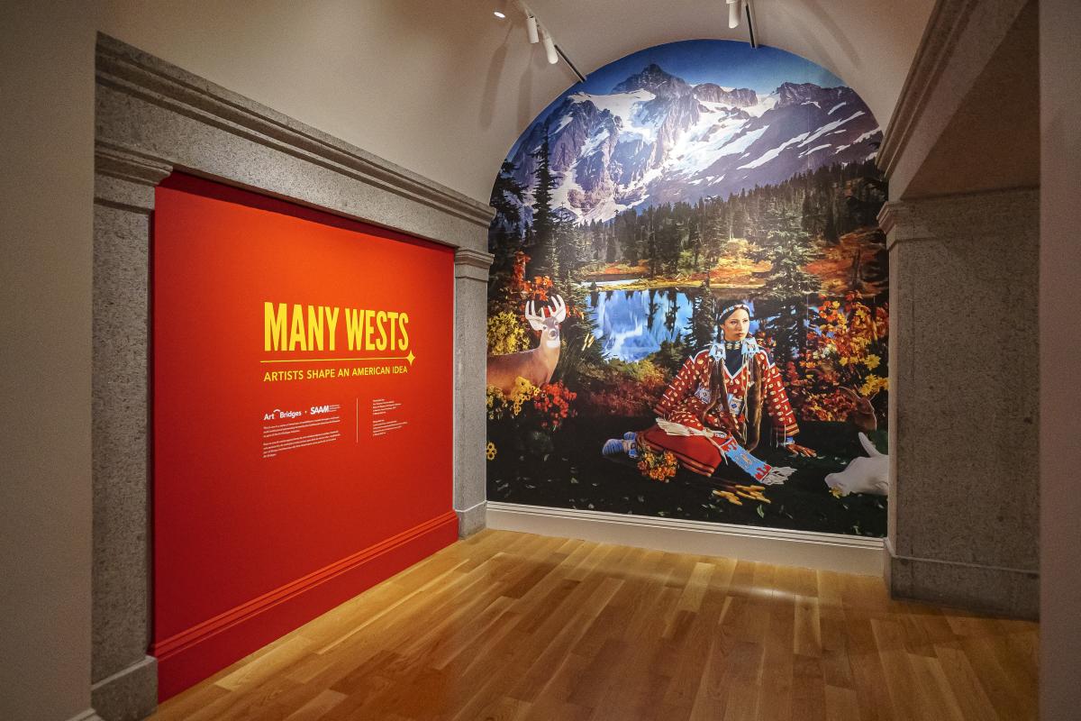 A mural at the end of a hallway. The left wall is painted deep red. The mural is of a painting of a Native woman sitting in front of a lake with snowy mountains in the distance.