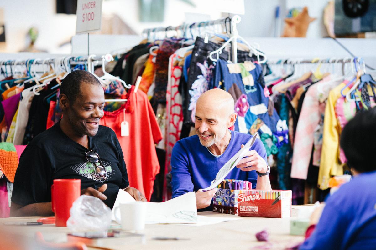 Two men sit at a long table with artist supplies such as markers and racks of clothing behind them.