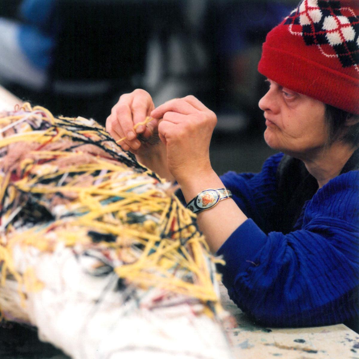 A woman in a red cap and a blue shirt works adds threads to a work of art