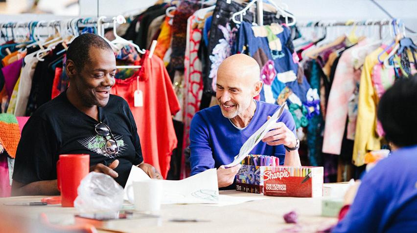 Three people sit at a table. Racks of clothes hang behind them.