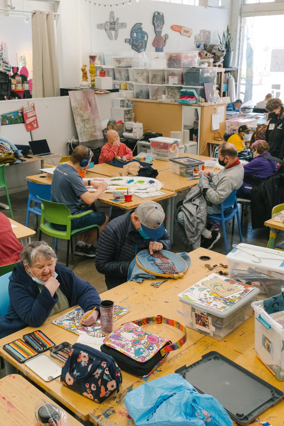 A room full of people working on various art projects in different media at long tables