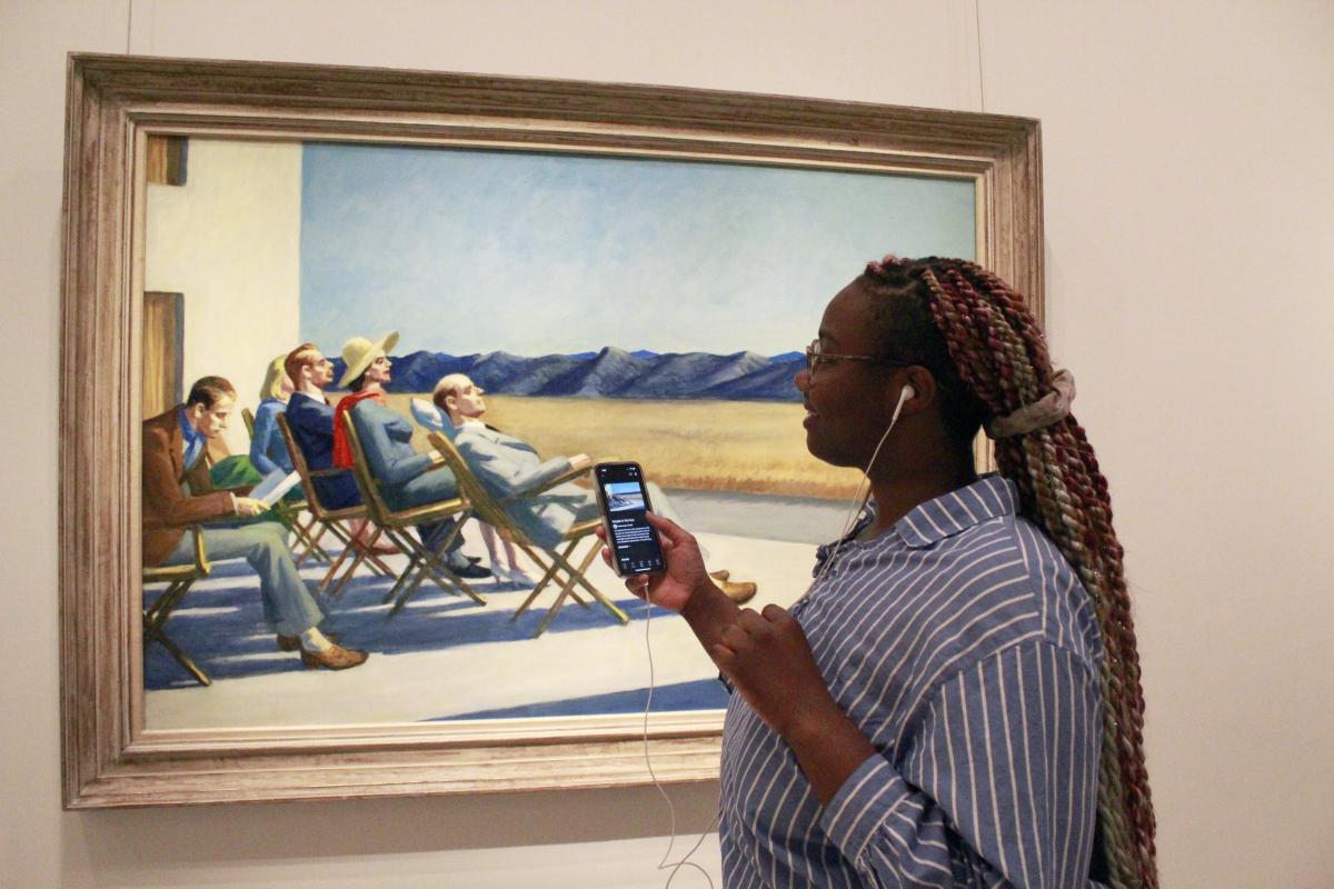 A photograph of a person standing in front of an artwork. She is wearing earbuds and holding her phone up to the work.