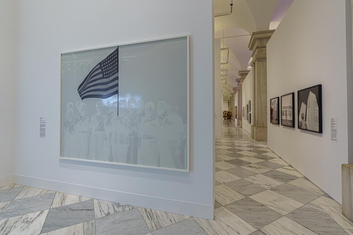 Installation view of a black and white print of a group of young boys gathered around a United States flag