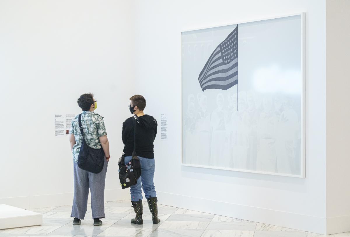 Two people stand in the corner of a room next to a large black and white print of a group of children with a United States flag