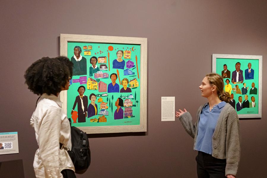 Two women look carefully at a colorful, intricate painting by William H. Johnson