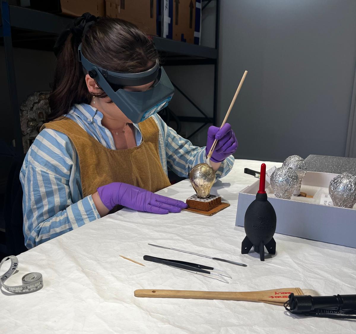 An art conservator, wearing magnifying glasses, brushes part of an artwork.