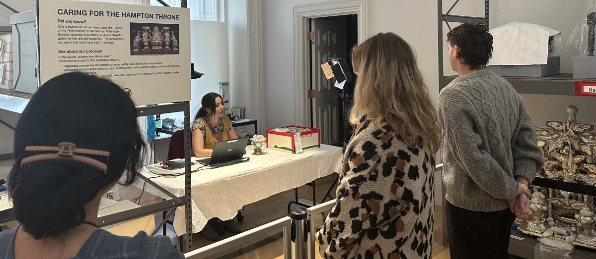 Visitors stand behind a rope. They are looking at a conservator who sits behind a table.