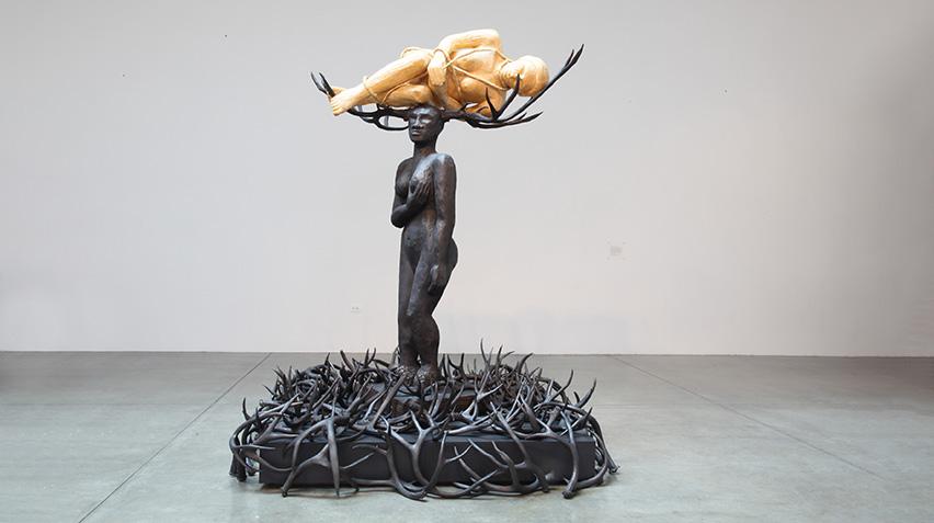 Sculpture of a woman. Massive antlers on her head cradle a delicate, translucent adult figure in a fetal position, like a creature preparing to emerge from its cocoon. Scattered on the ground below are antlers.