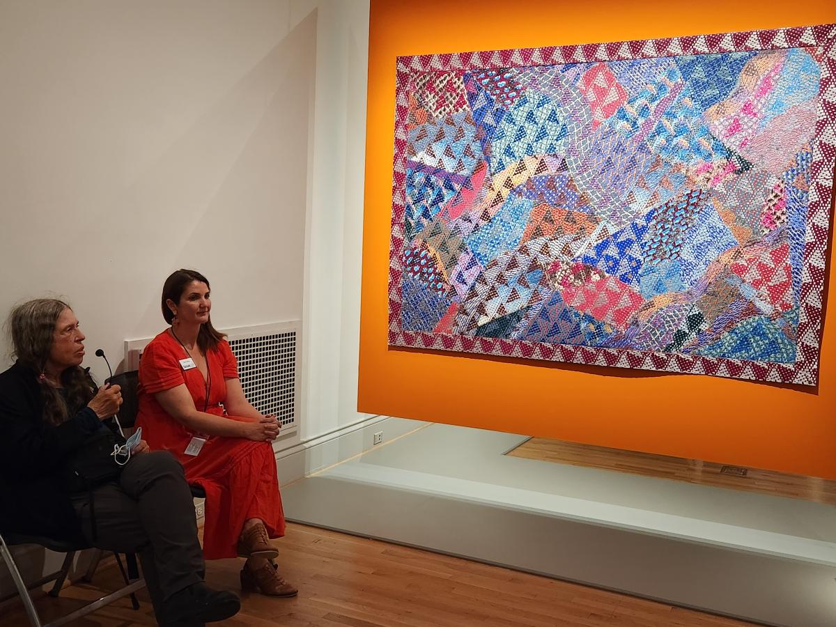 Two women sitting in a gallery looking at a work of fiber art hanging on the wall.