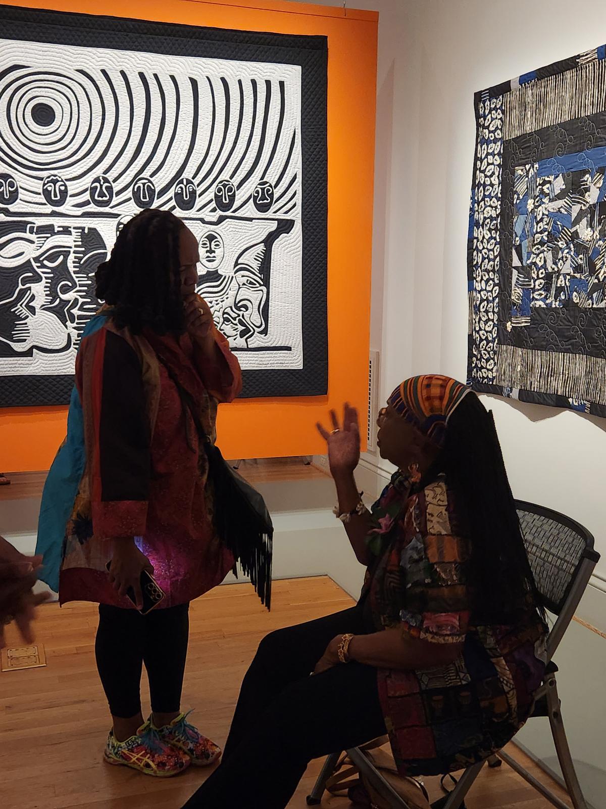 A person sitting in a chair looks up at a person in front of her. Behind them, a black and white artwork hangs on an orange wall.