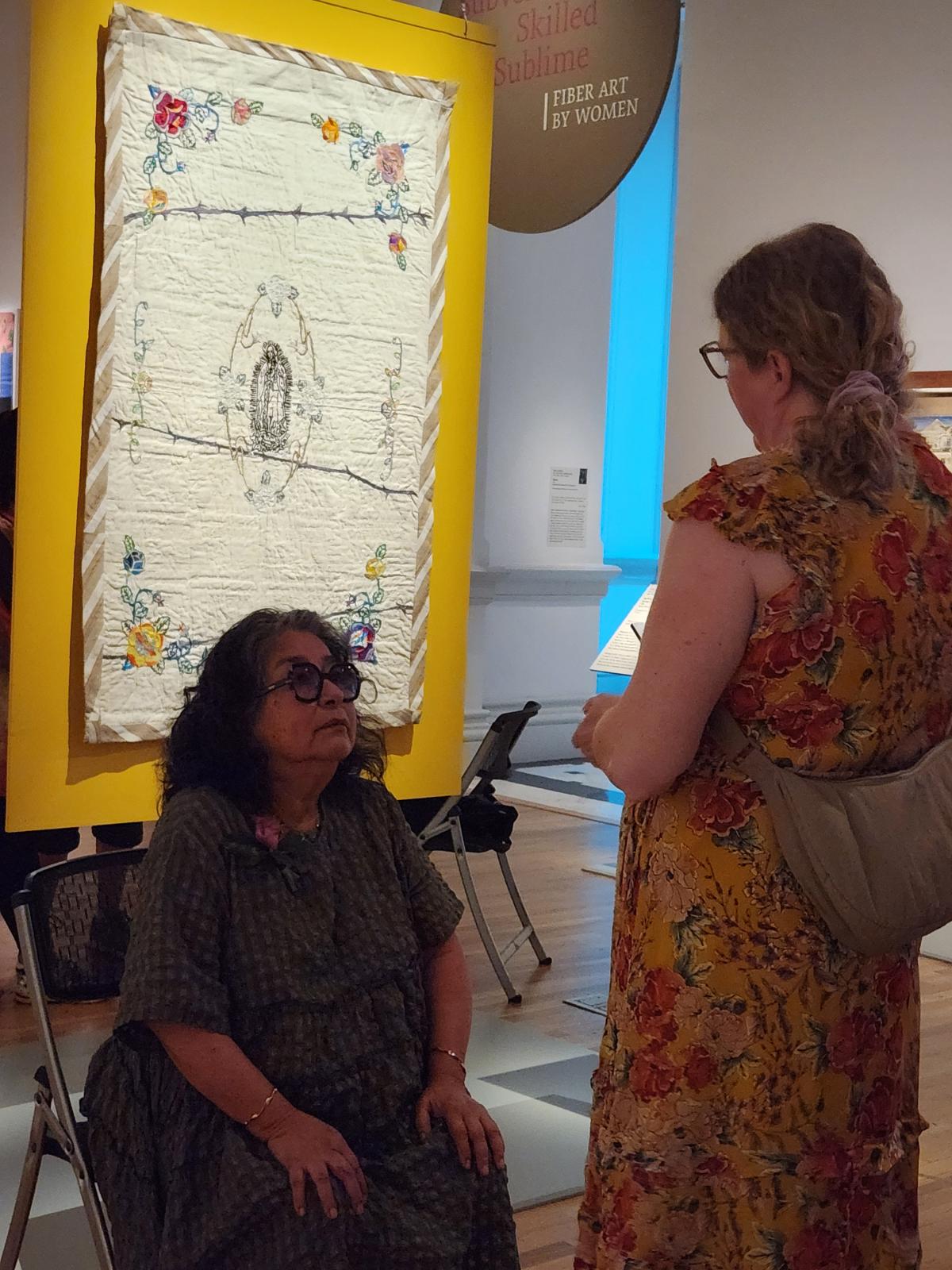 A person sitting in a chair looks up at a person in front of her. Behind them, an artwork hangs on a yellow wall.