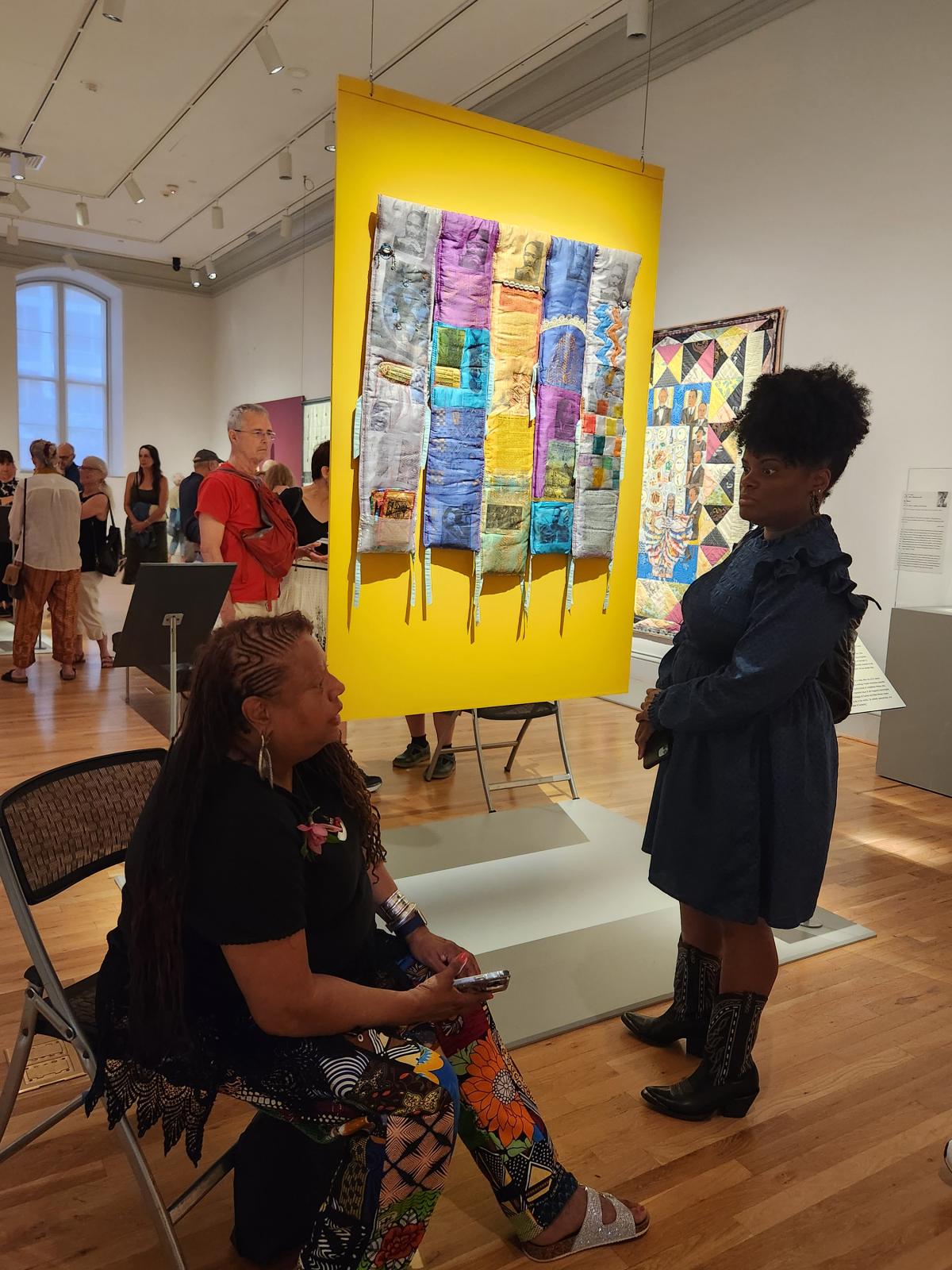 A person sitting in a chair looks up at a person in front of her. Behind them, an artwork hangs on a yellow wall.