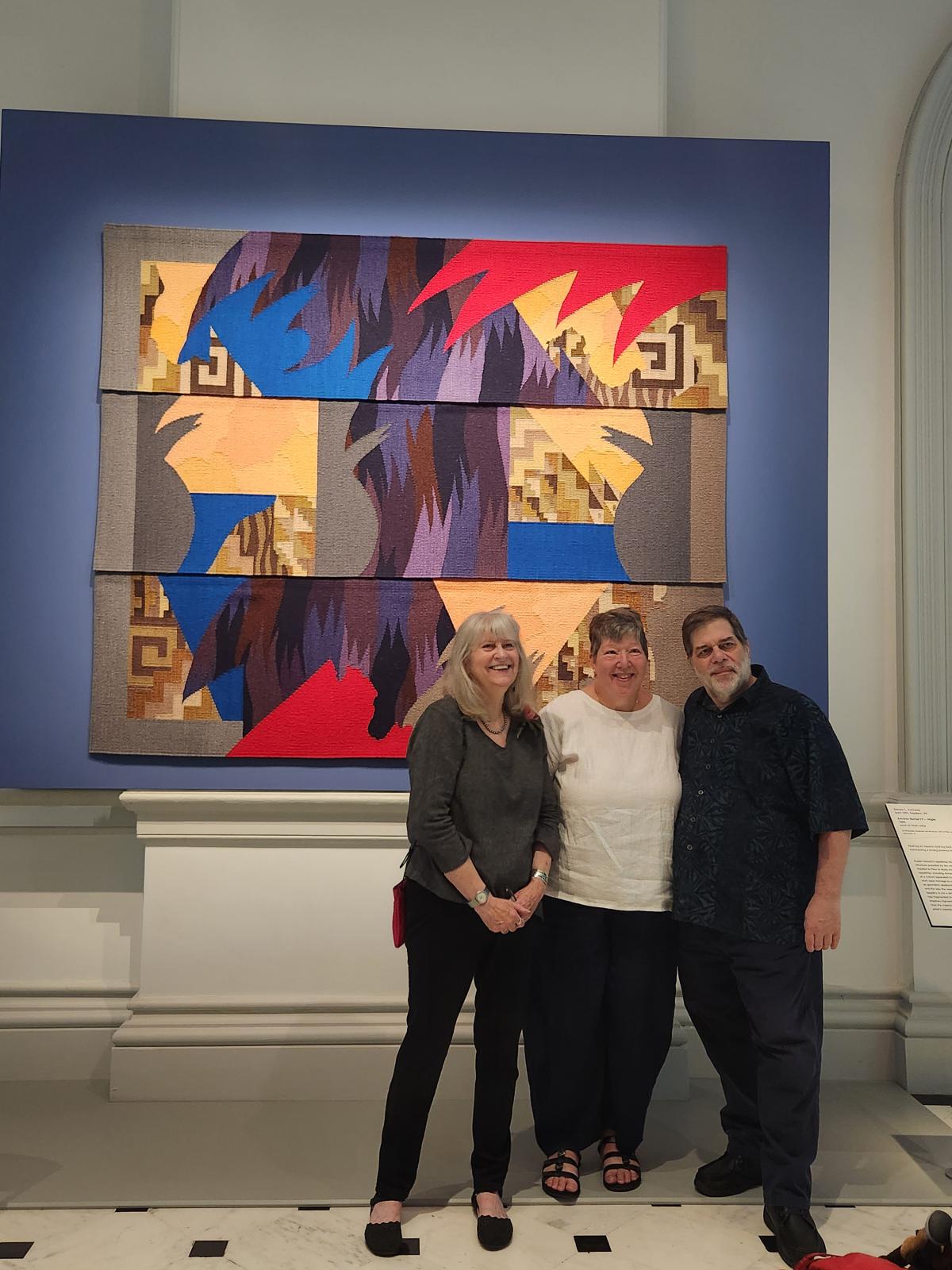 Two women and a man pose for a photo in front of an abstract artwork that hangs on a blue wall.