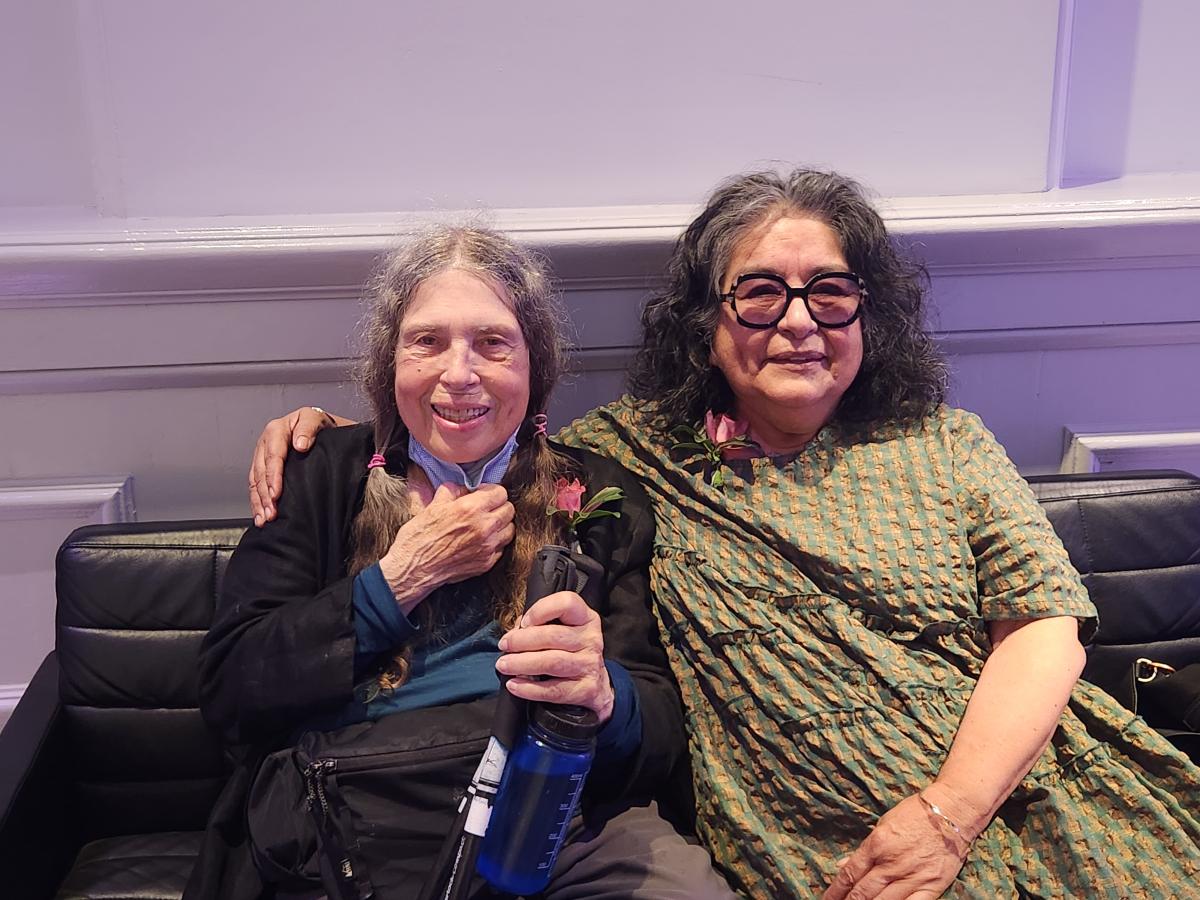 A woman sits on a bench with her arm around another woman. They are smiling for the camera.