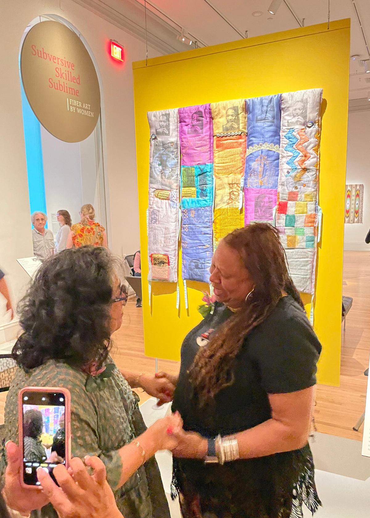 Two women smile at each other. Behind them, an artwork hangs on a bright yellow wall.