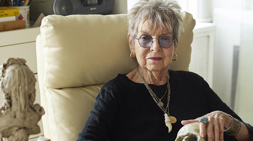 Audrey Flack sits in a chair. Her hand is on a skull.