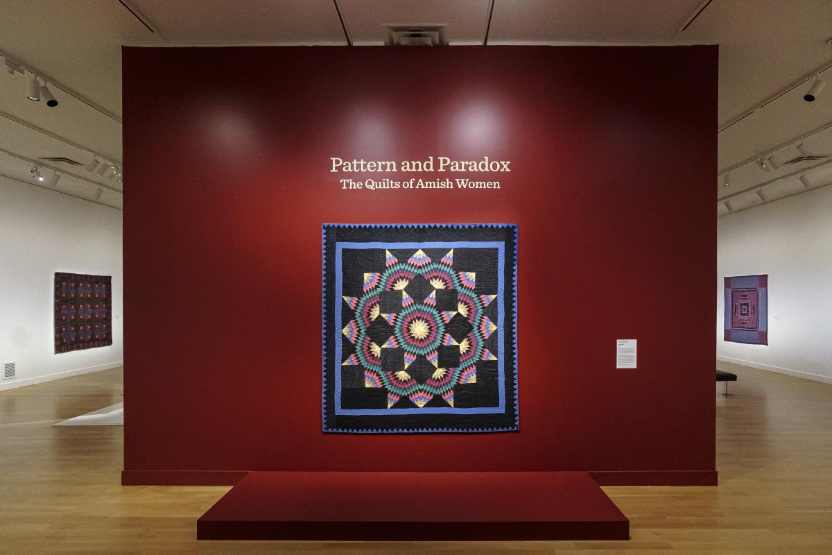 Dark red wall with a quilt hanging in the center. Above it is the title "Pattern and Paradox: The Quilts of Amish Women."