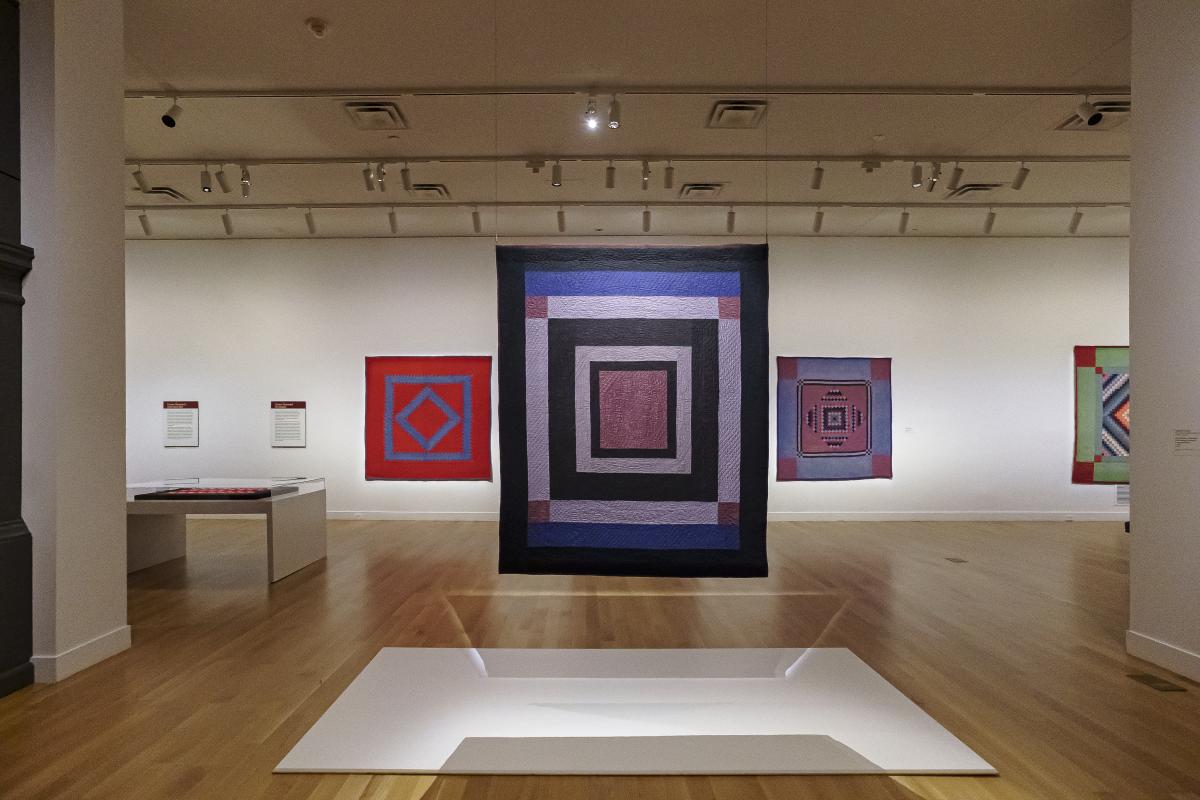 A quilt with large, concentric squares hang in a gallery. Quilts are mounted on the wall behind it.