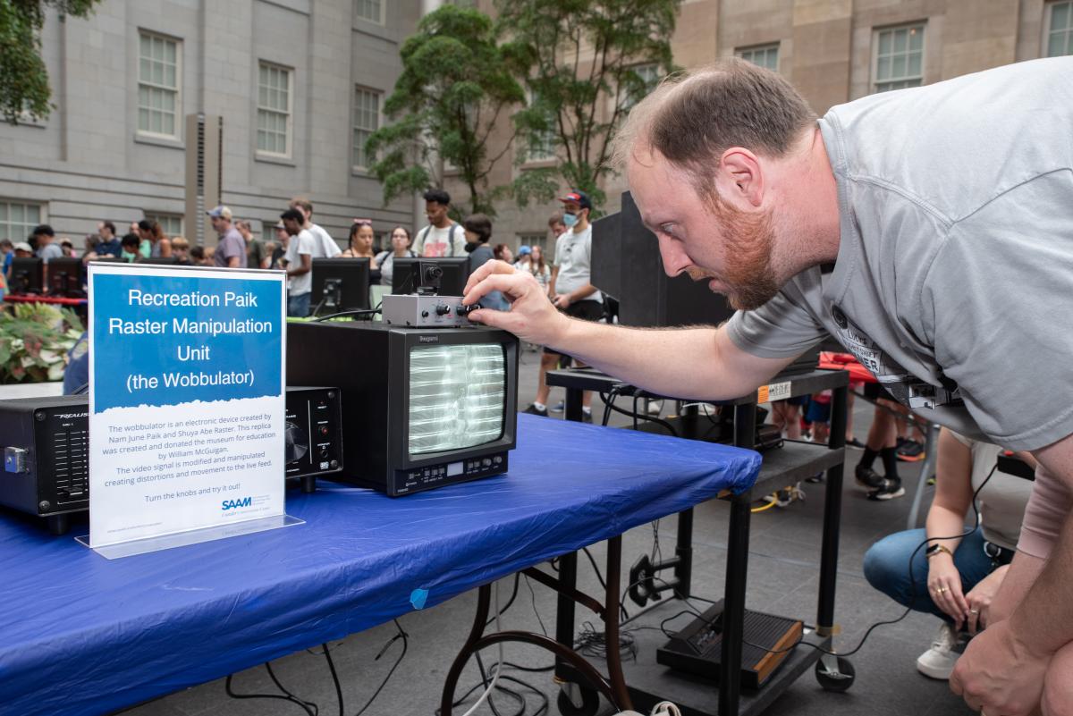 A man turns the nobs of a box sitting atop a small black and white tv monitor.