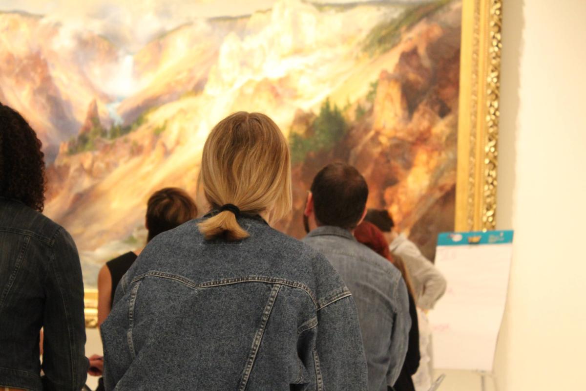 A woman with her back to the camera gazes at a large landscape painting in a gold frame