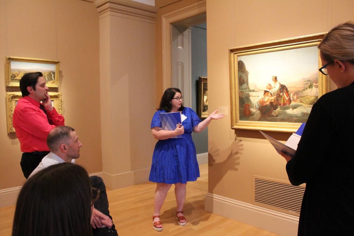 A teacher in a blue dress looks carefully at a 19th century painting in a gold frame