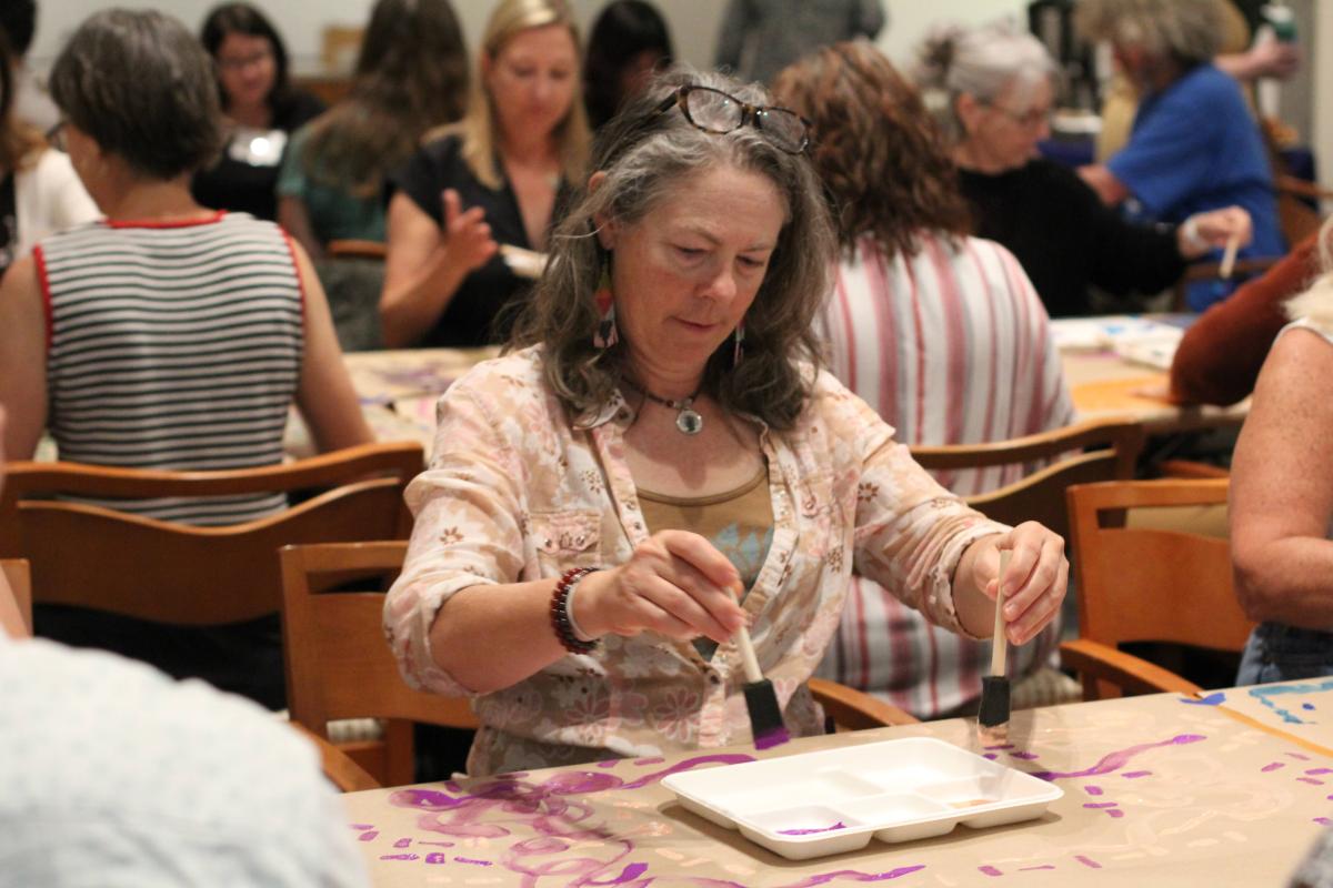 A teacher practices art making at a long table with paints and paper in front of her.