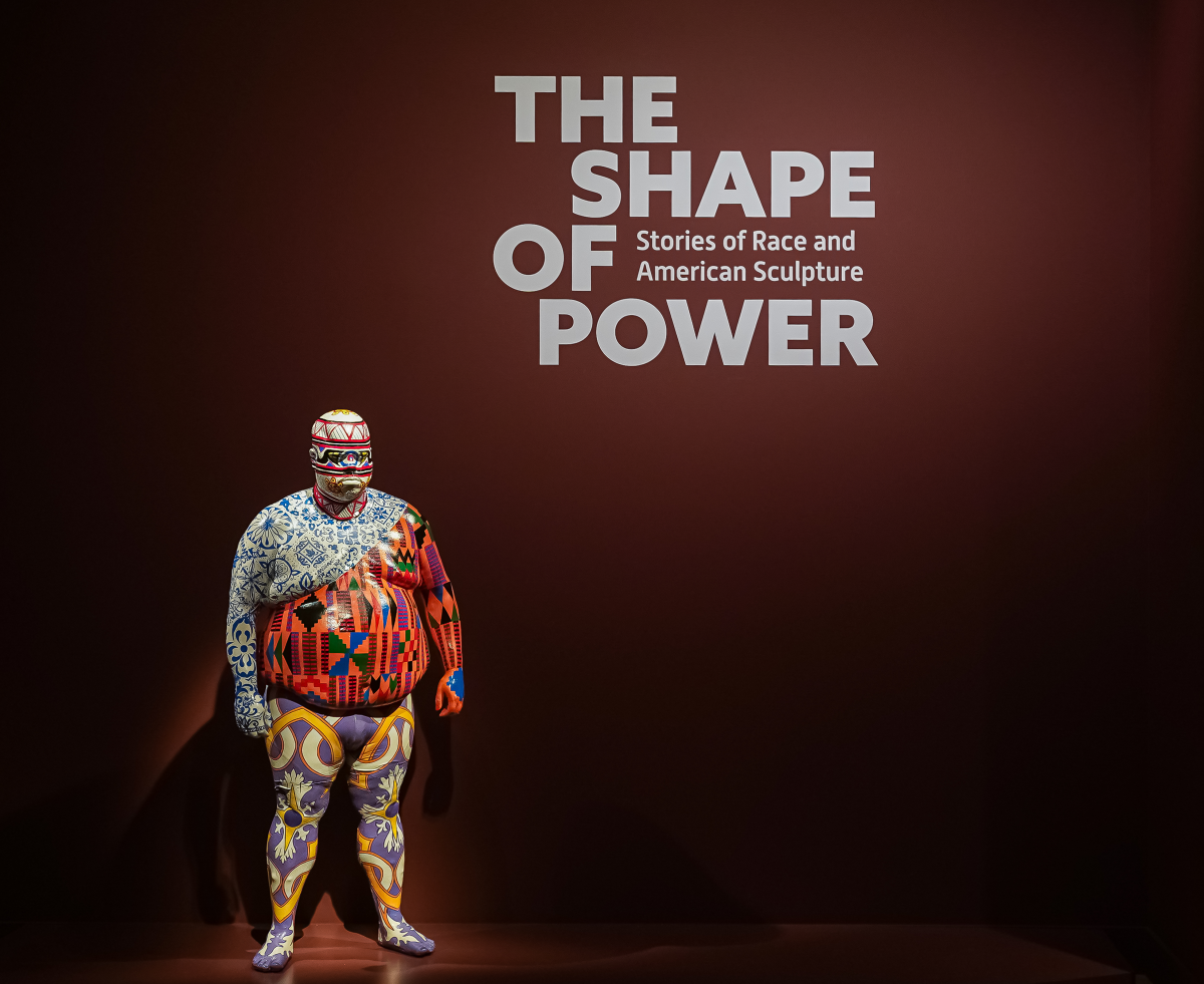 Sculpture of a person completely covered with multiple colorful, intricate patterns standing against a dark red wall with the exhibition title "The Shape of Power: Stories of Race and American Sculpture."