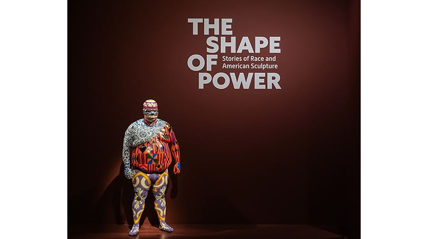 Sculpture of a person completely covered with multiple colorful, intricate patterns standing against a dark red wall with the exhibition title "The Shape of Power: Stories of Race and American Sculpture."
