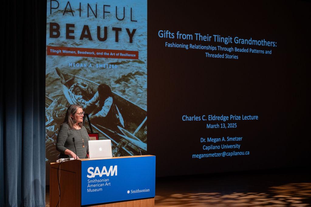 A speaker stands at a podium during a lecture. A large screen behind them displays the talk title,