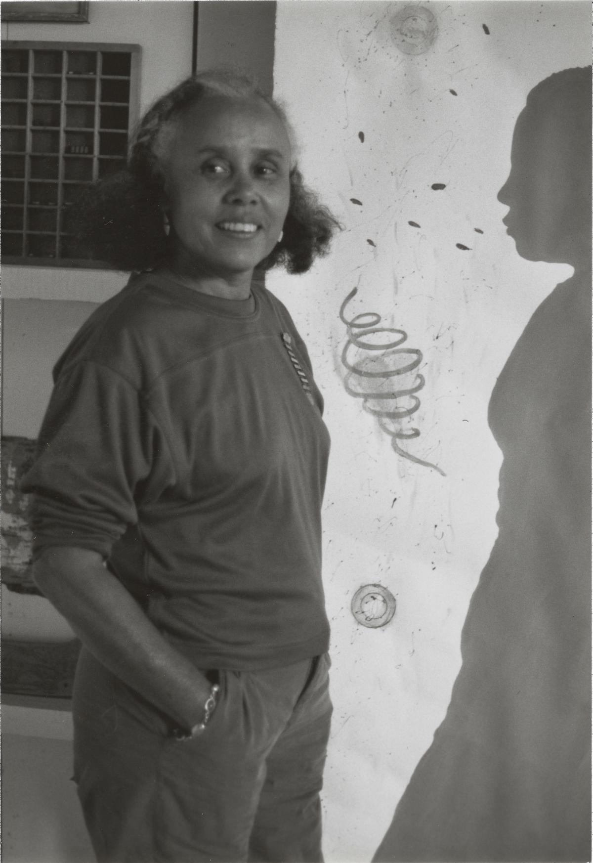  A black-and-white photograph of a woman with short hair smiling, wearing a long-sleeved top and pants. Her hands are in her pockets, and she is turning to face the camera. Behind her is wall art in various shapes, including the shadow of a figure. 