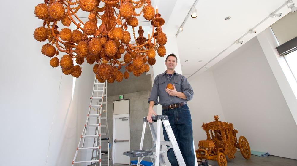 Artist Timothy Horn standing on ladder next to his large chandelier sculpture made of rock sugar