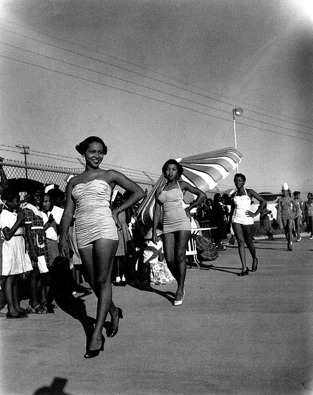 A photo of women in a beauty pageant with observers on the street behind them