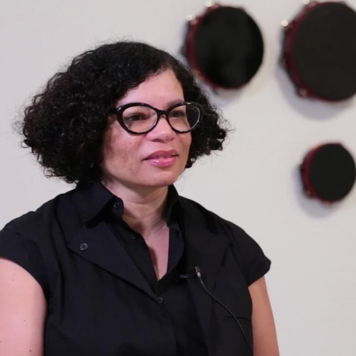 A photograph of a woman sitting down with artwork on the wall behind her