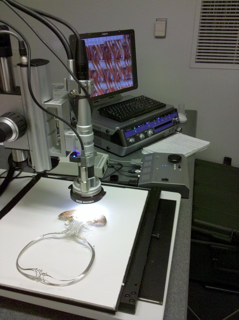 A silver choker on a white sheet is shown under conservation equipment that is beaming light on it