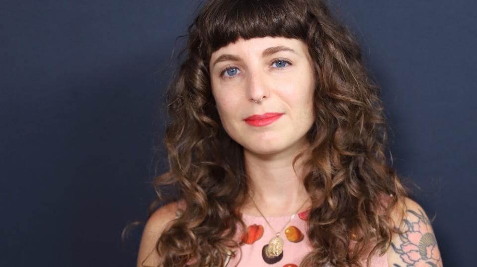 Sasha, a white femme with long brown curly hair, blunt bangs, red lipstick and a pink top.